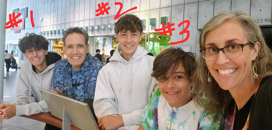 A smiling family of five sitting at a table in a casual setting. From left to right: a teenage boy labeled #1 with curly hair and wearing a grey hoodie, a man in a blue patterned shirt, a teenage boy labeled #2 wearing a light grey hoodie, a younger boy labeled #3 with wavy hair in a colorful tie-dye shirt, and a woman with glasses and wavy hair on the far right, smiling brightly.