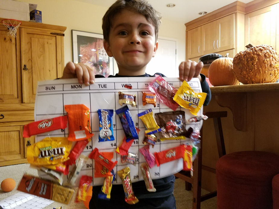 A young boy holds up a calendar decorated with various candy pieces attached to each date, creating a fun candy countdown or reward calendar. In the background, a pumpkin sits on the kitchen counter, adding a seasonal touch.