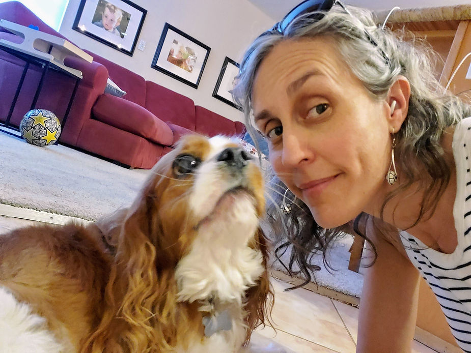 A woman with sunglasses resting on her head smiles while posing next to a Cavalier King Charles Spaniel. The dog looks up at her affectionately. They are in a comfortable living room with a red couch, framed photos on the wall, and a soccer ball in the background. The atmosphere feels warm and relaxed, as they share a quiet moment together.