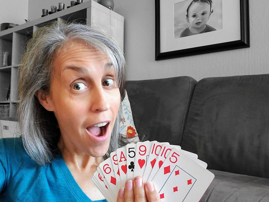 A woman with gray hair is holding a fan of playing cards, looking surprised or excited. She is sitting on a couch, and there is a framed photo of a child in the background.