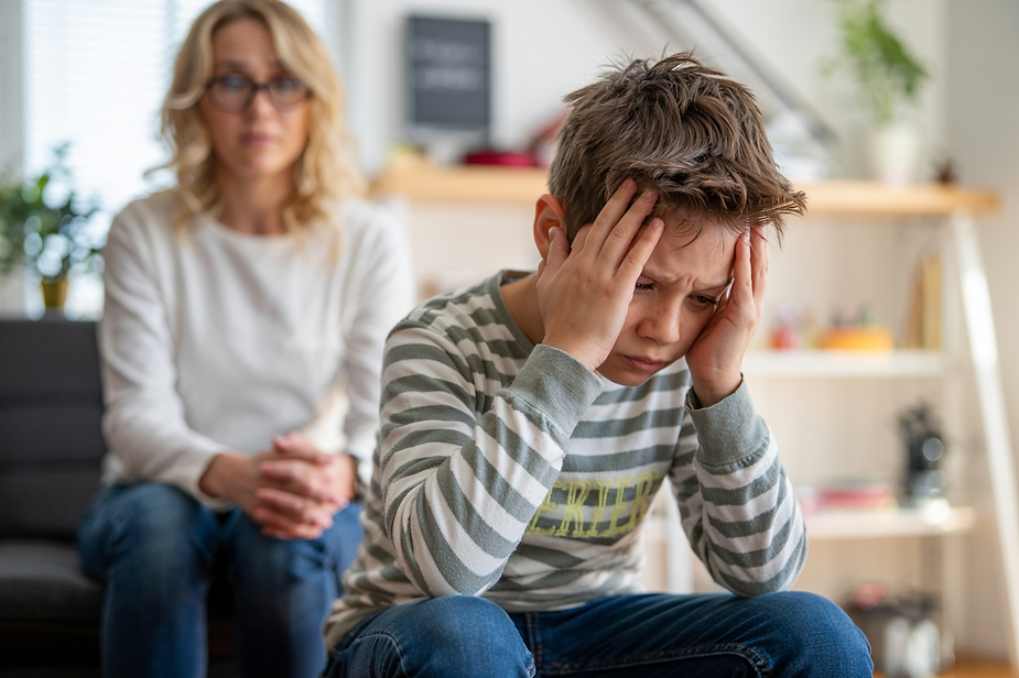 A frustrated boy holds his head while his mom watches with concern in the background.
